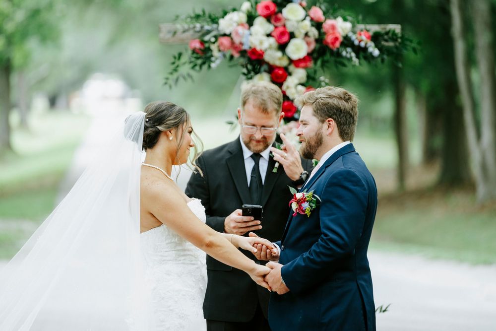 Outdoor wedding ceremony with bride, groom, and officiant under floral arch of red and pink blooms on tree-lined path.