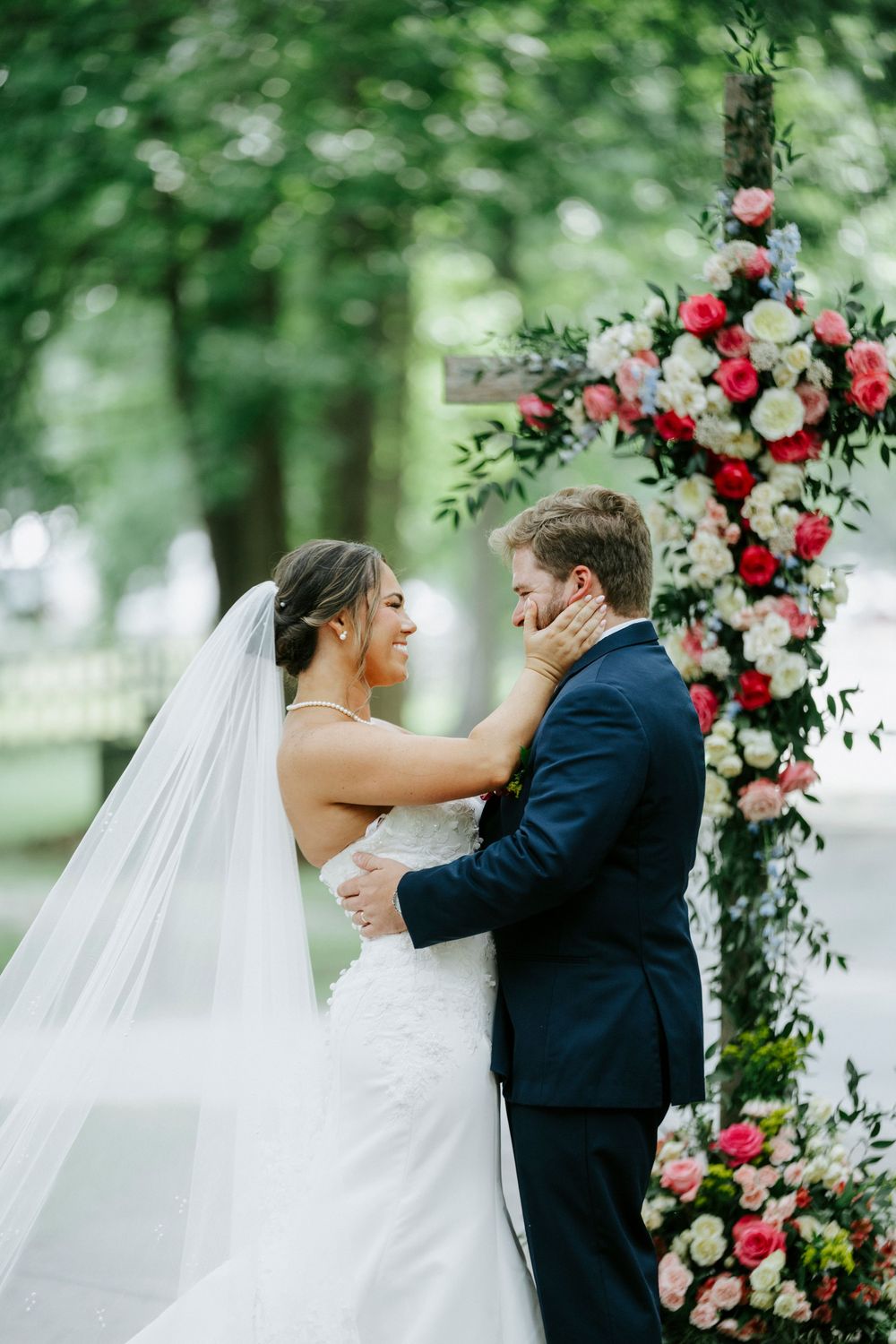 Bride in lace gown tenderly cups grooms face near floral arch of pink and white roses.