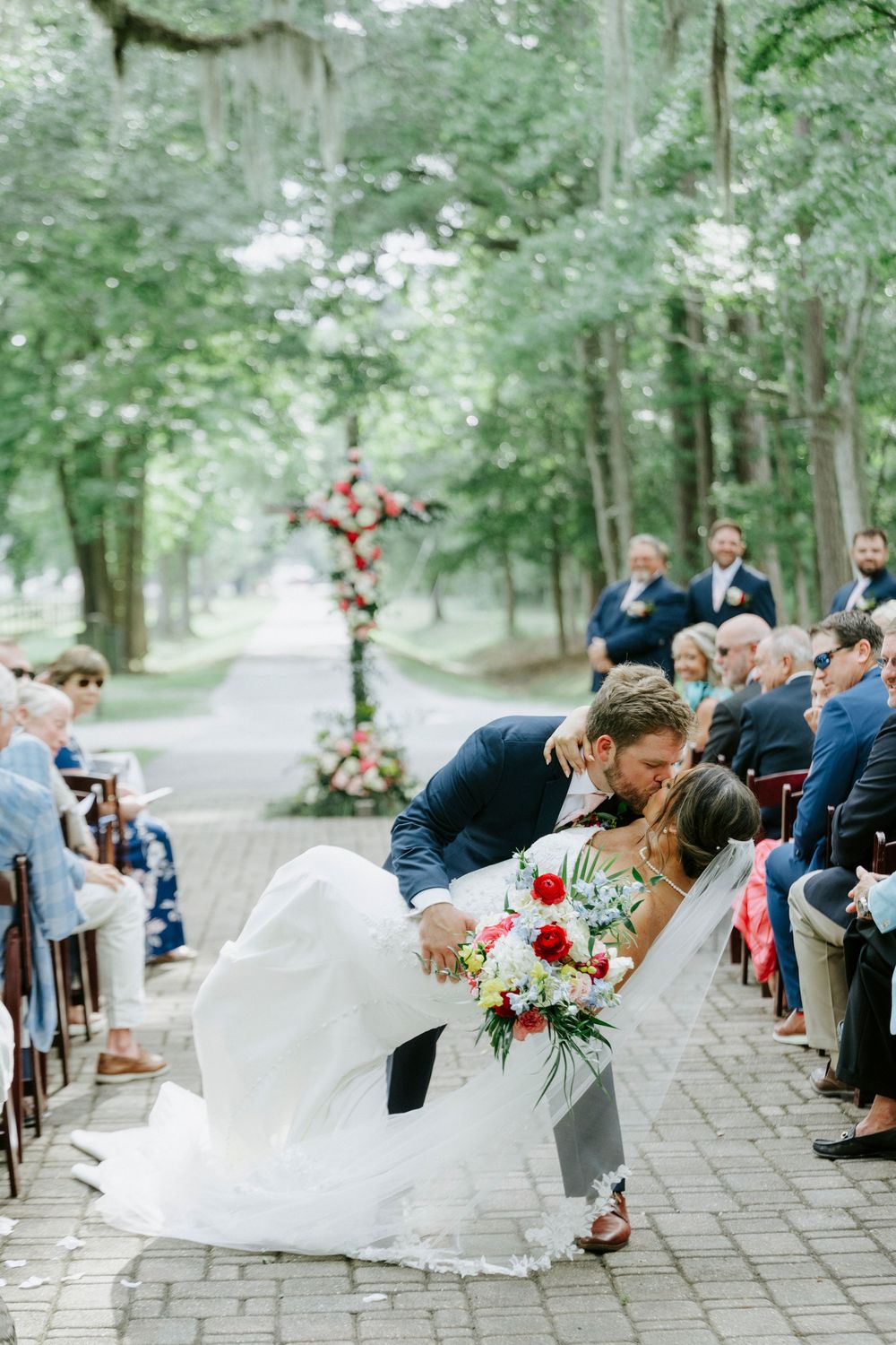 Groom dips bride for romantic kiss at end of aisle during outdoor wedding ceremony lined with seated guests.