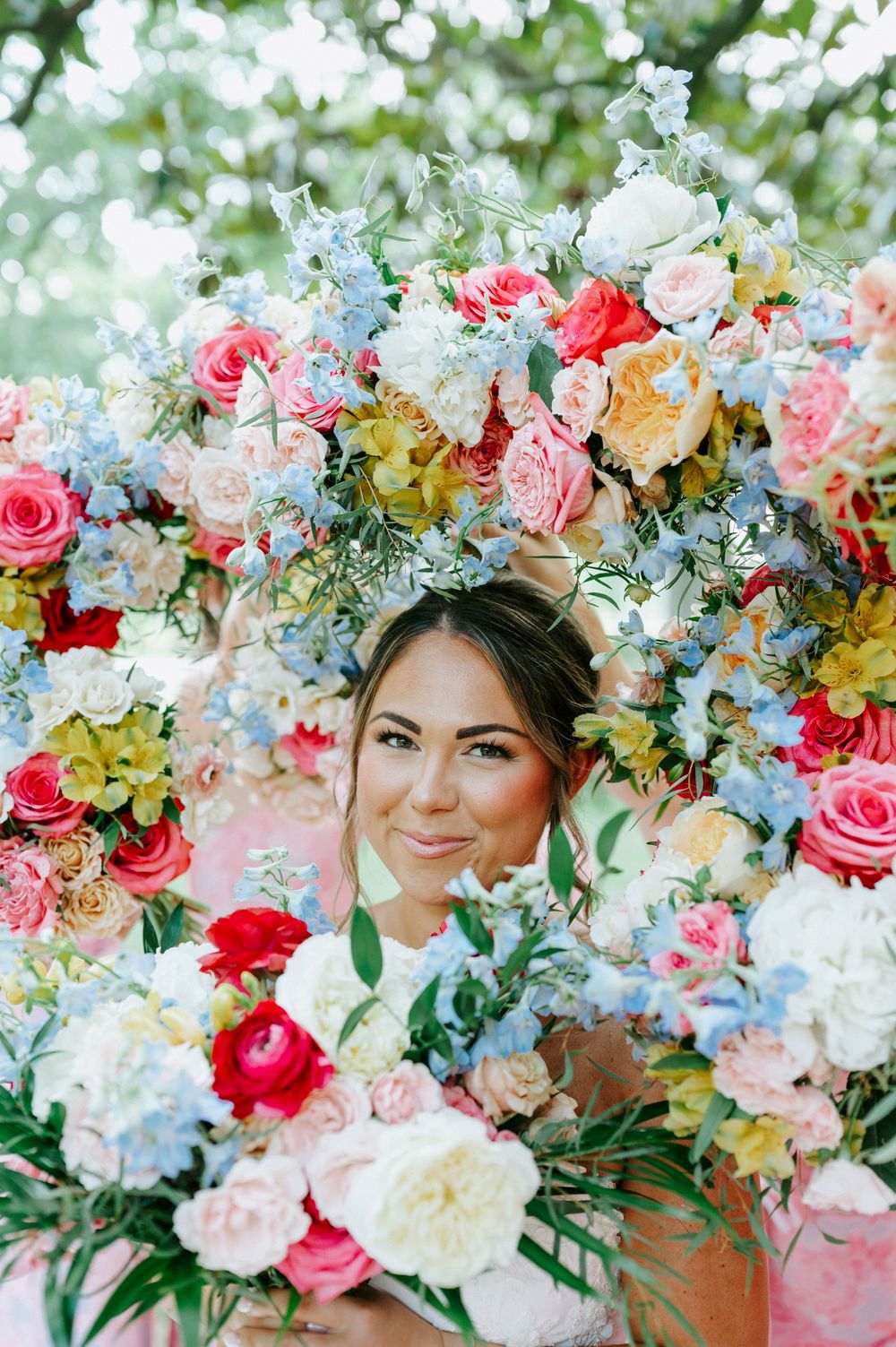 Smiling bride peeks through a stunning circular floral arrangement of pink, blue, and white roses and mixed blooms.