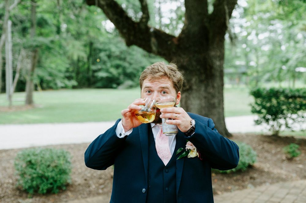 Groom in navy suit drinking whiskey outdoors near large oak tree at wedding reception.