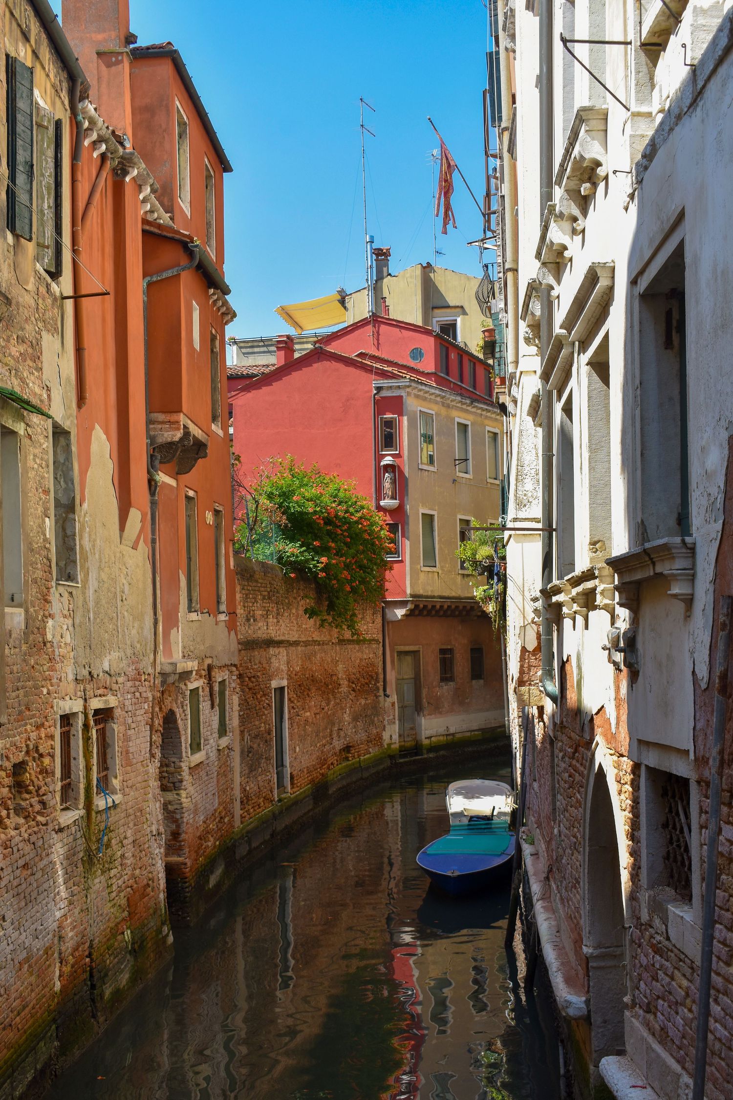 Narrow Venice canal with colorful historic buildings, a moored boat, and reflections on calm water under a bright blue sky.