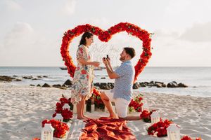 Man proposes to woman on beach with red heart floral arch and rose petals at sunset.