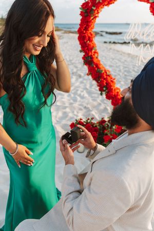 Man kneeling on beach proposing to woman in green dress with red floral arch backdrop at sunset.
