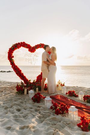 Couple kissing under a red rose heart arch with Will You Marry Me sign on a romantic beach at sunset.