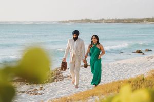 Couple walking hand in hand on a tropical beach, man in beige suit, woman in green dress, ocean waves behind them.