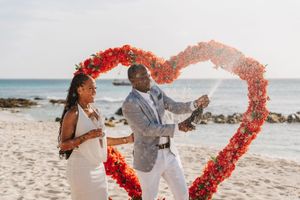 Couple celebrating on beach under red heart floral arch, man popping champagne, woman in white dress, ocean backdrop.