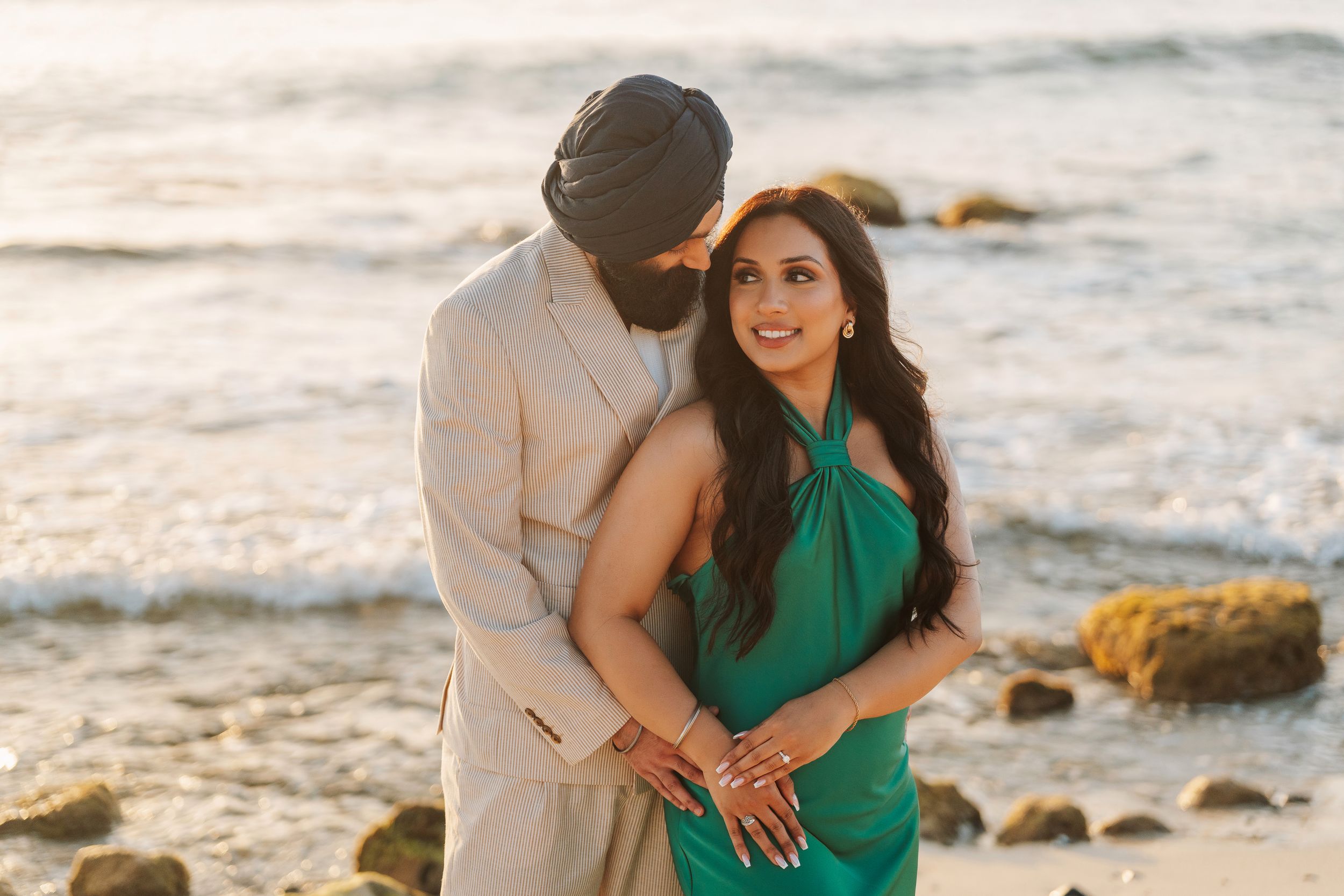 Couple in elegant attire embracing on a rocky beach at golden hour, woman in green dress, man in beige suit with turban.
