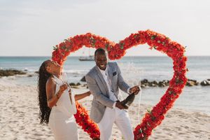 Joyful couple celebrating engagement with champagne in front of a red rose heart arch on a sunny beach.