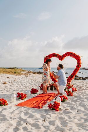 Man proposes to woman under red heart-shaped floral arch on white sand beach with red roses and candles.