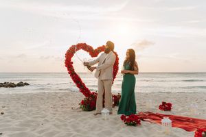 Couple celebrating beach proposal at sunset with red heart floral arch, rose petals, and candles on sandy shore.