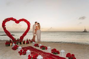Couple kissing under a red rose heart arch on a beach at sunset with candles and rose petals.