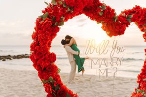 Couple embracing under a red floral heart arch on the beach with a Will You Marry Me neon sign at sunset.