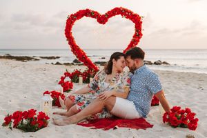 Romantic couple sitting on beach with red heart floral arch, roses, and candles during a Valentine's Day proposal.