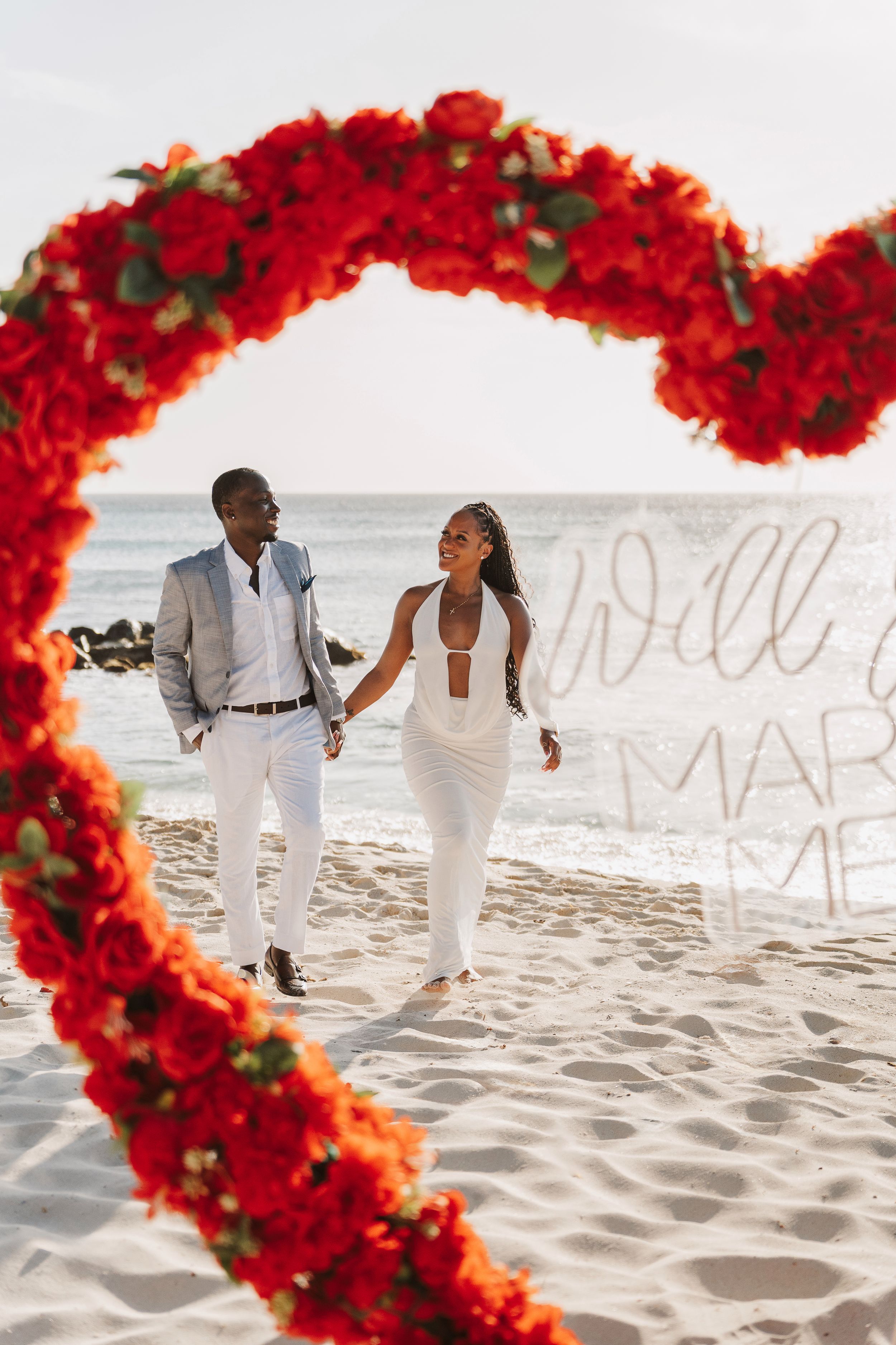 Happy couple holding hands walking through a red floral heart arch on a sandy beach with marry me sign.