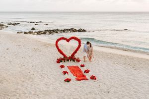 Couple embracing on a white sand beach beside a large red heart rose arch with flower petals for a romantic proposal.