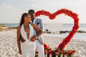Couple embracing on beach in front of red floral heart arch, woman in white dress, man in gray suit.