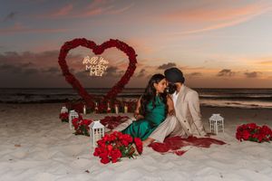 Couple shares romantic beach proposal at sunset with red rose heart arch and Will You Marry Me neon sign.