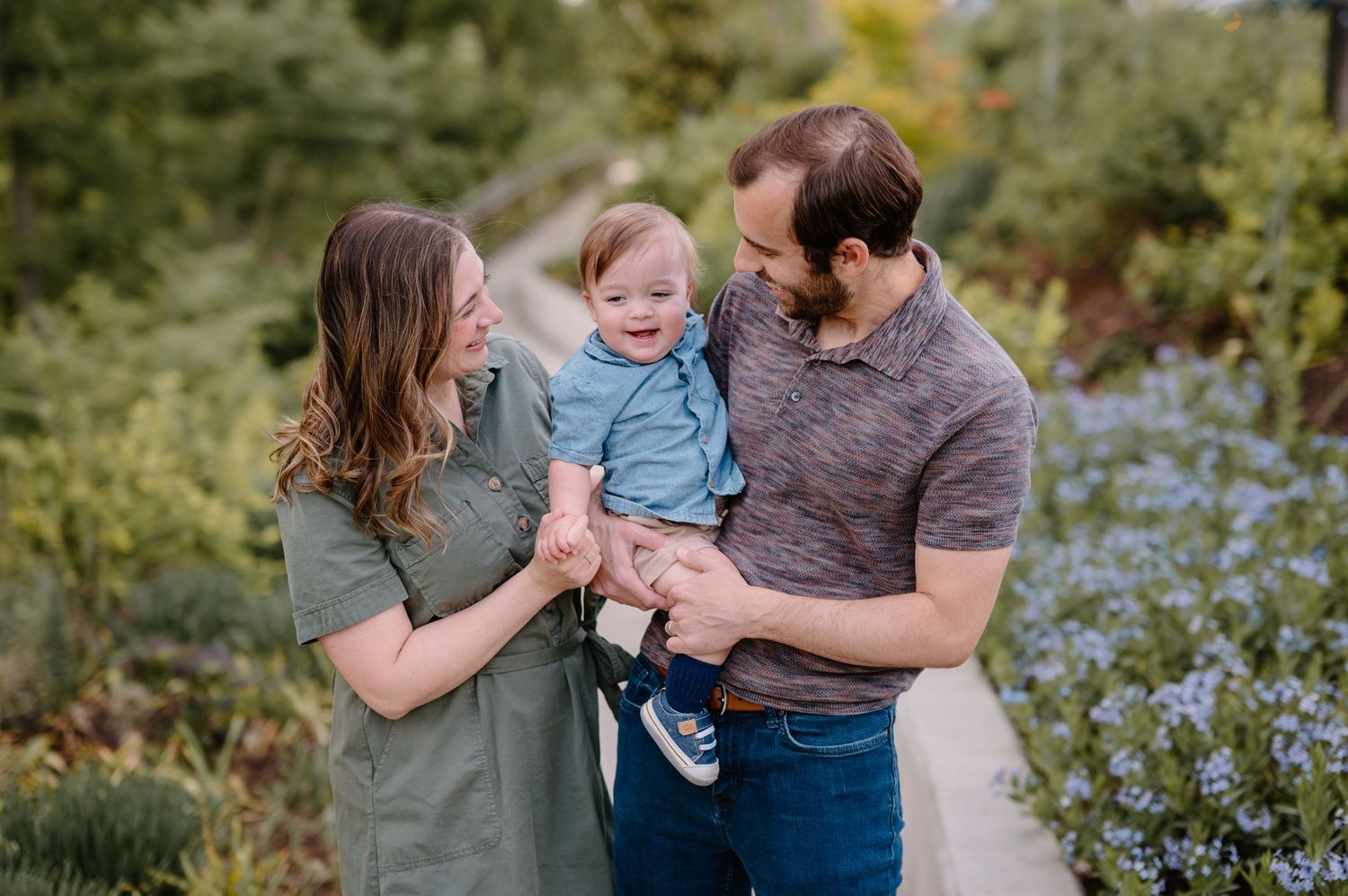 Happy family with mother, father, and baby boy smiling outdoors among blue wildflowers on a garden path.
