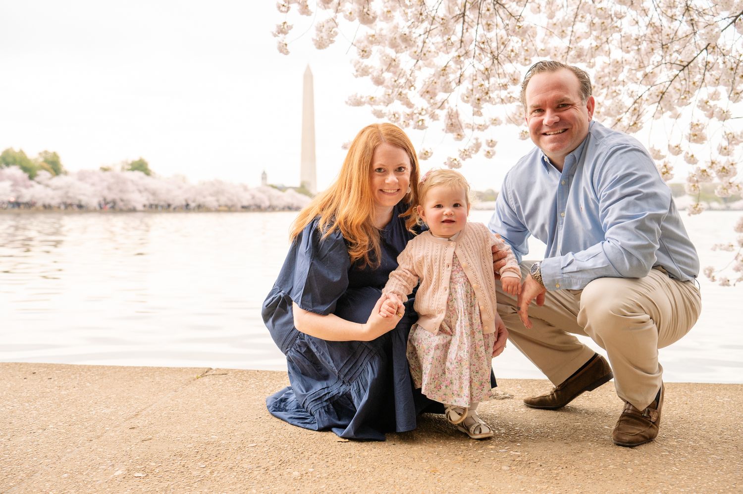 Family of three kneel, posed, by Tidal Basin in Washington DC, with cherry blossoms and Washington Monument in background.