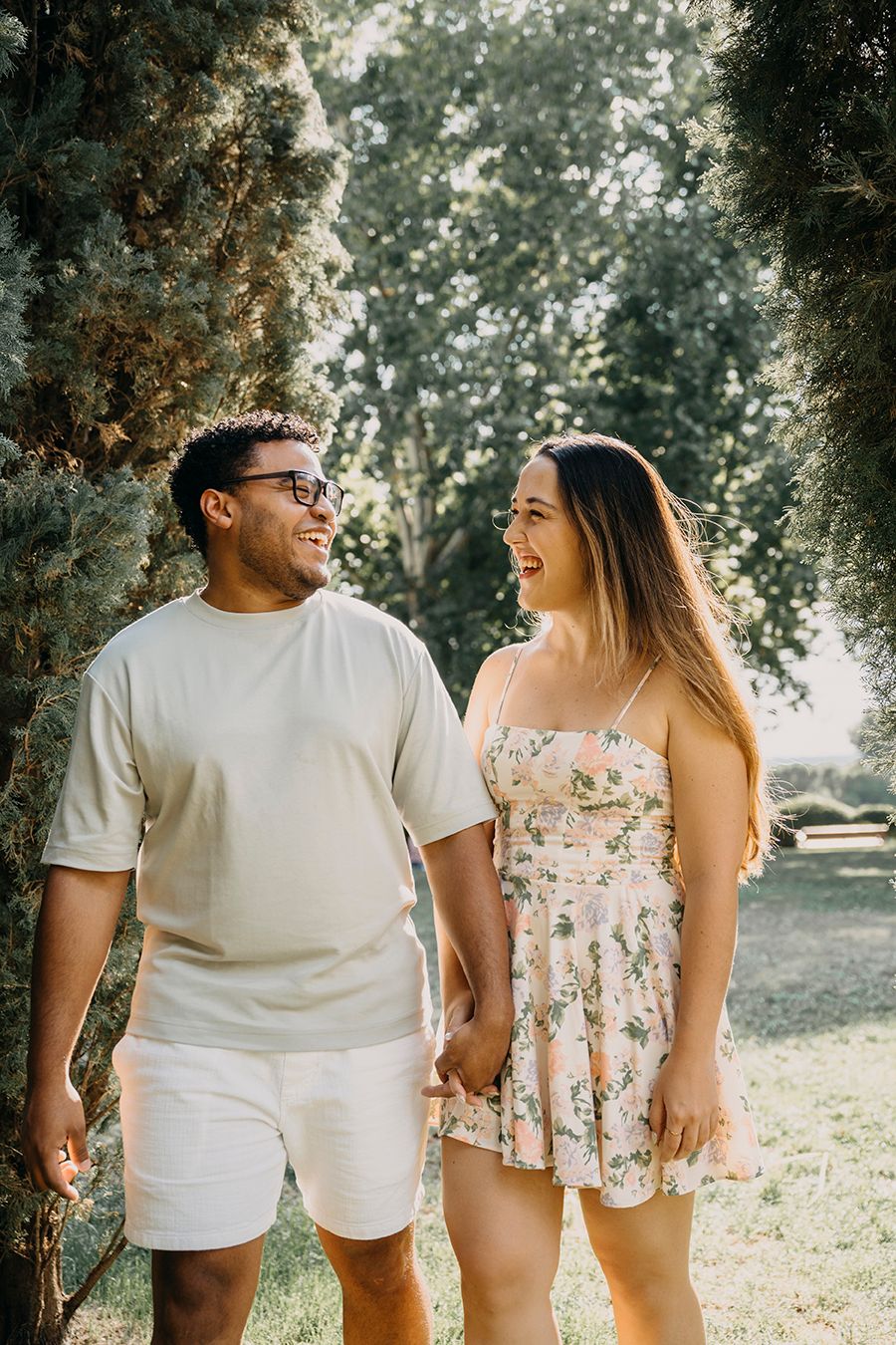 A smiling couple holds hands and walks through a sunny park, man in white outfit and woman in floral dress.