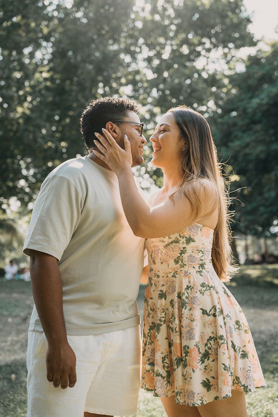 A couple sharing a romantic moment outdoors, woman in floral dress touching mans face, surrounded by lush green trees.