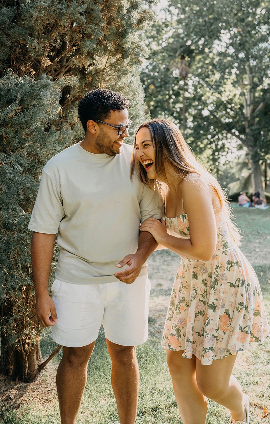 A laughing couple in casual summer outfits sharing a joyful moment together in a sunny park setting.