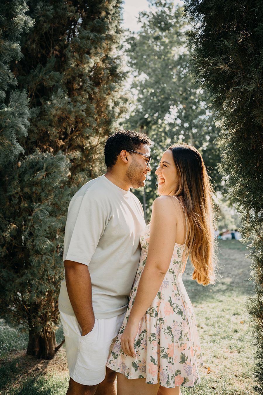 A romantic couple gazes at each other among tall evergreen trees in a sunlit park setting.