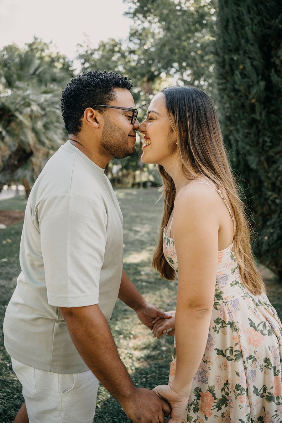 Couple holding hands and sharing a romantic moment nose-to-nose, smiling outdoors in a lush green park setting.
