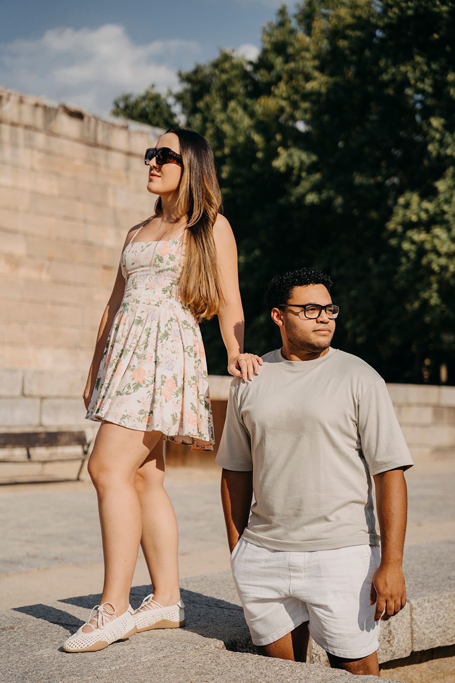 A couple holding hands outdoors, woman in floral dress with sunglasses, man in gray t-shirt and shorts.