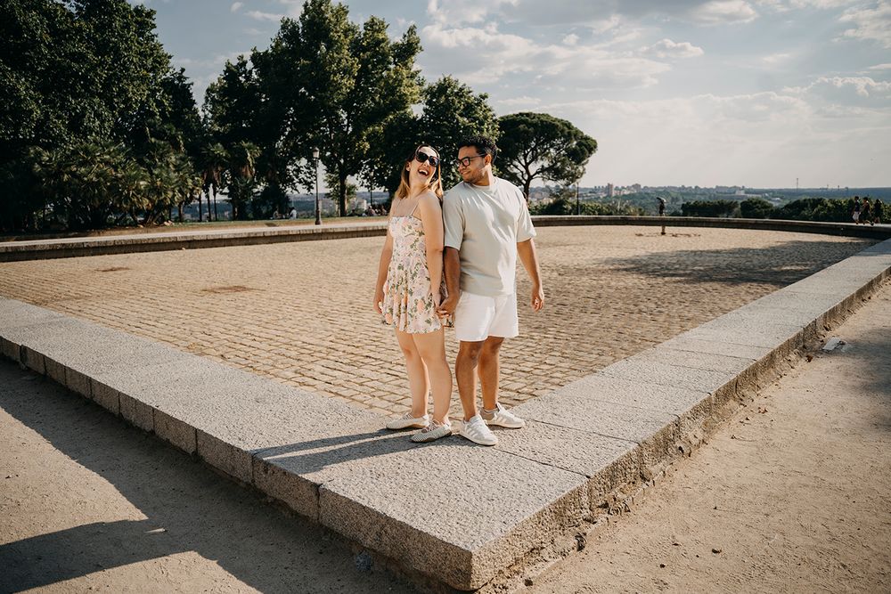 Couple posing together on a stone terrace with sandy ground and lush green trees in the background.