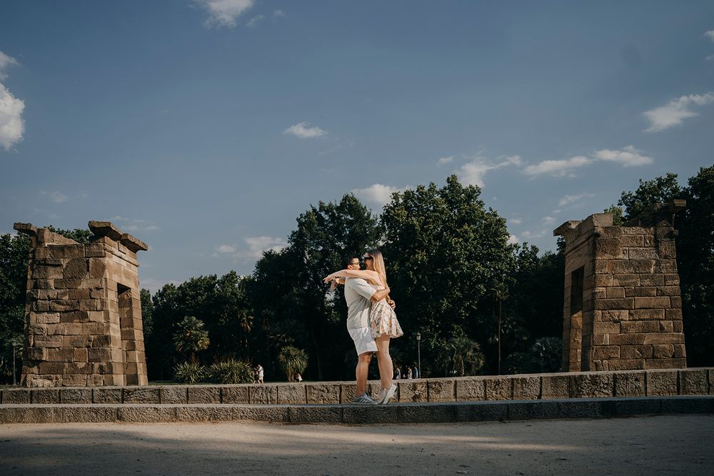 A romantic couple embracing on a stone wall in a sunny park with blue skies and green trees.