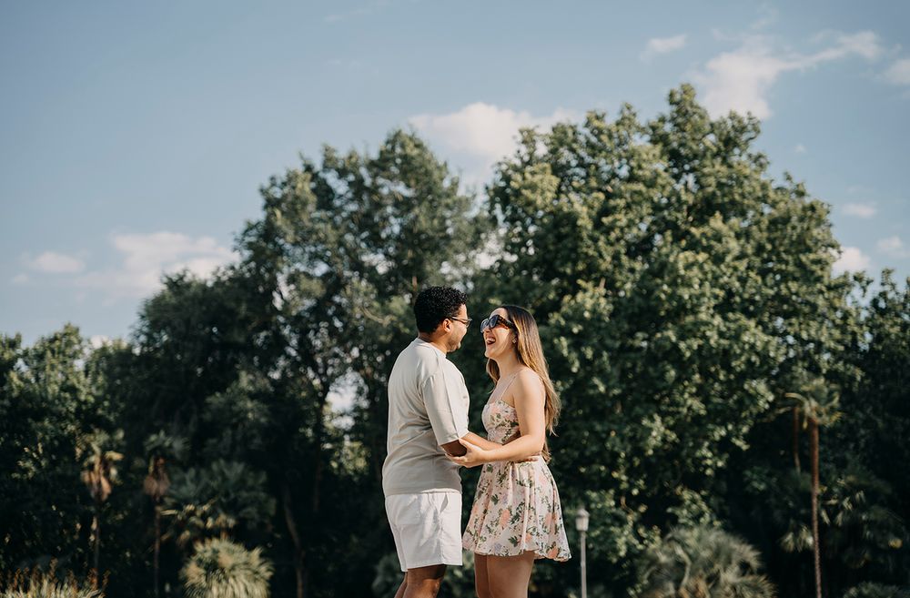 A couple stands facing each other in a lush green park, sharing a romantic moment under a partly cloudy sky.