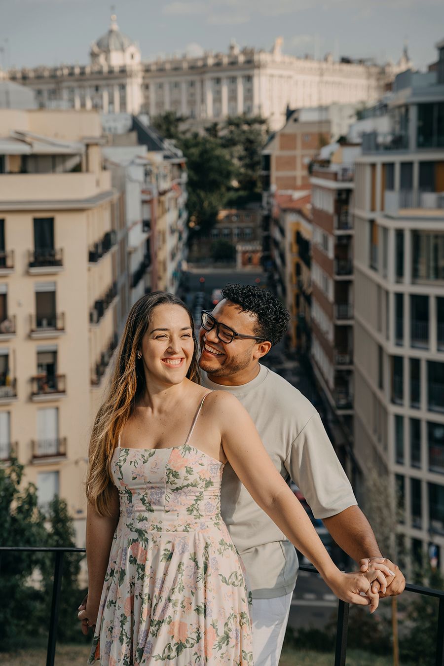 Happy couple holding hands on a scenic Madrid balcony, woman in floral dress, city buildings and palace in background.