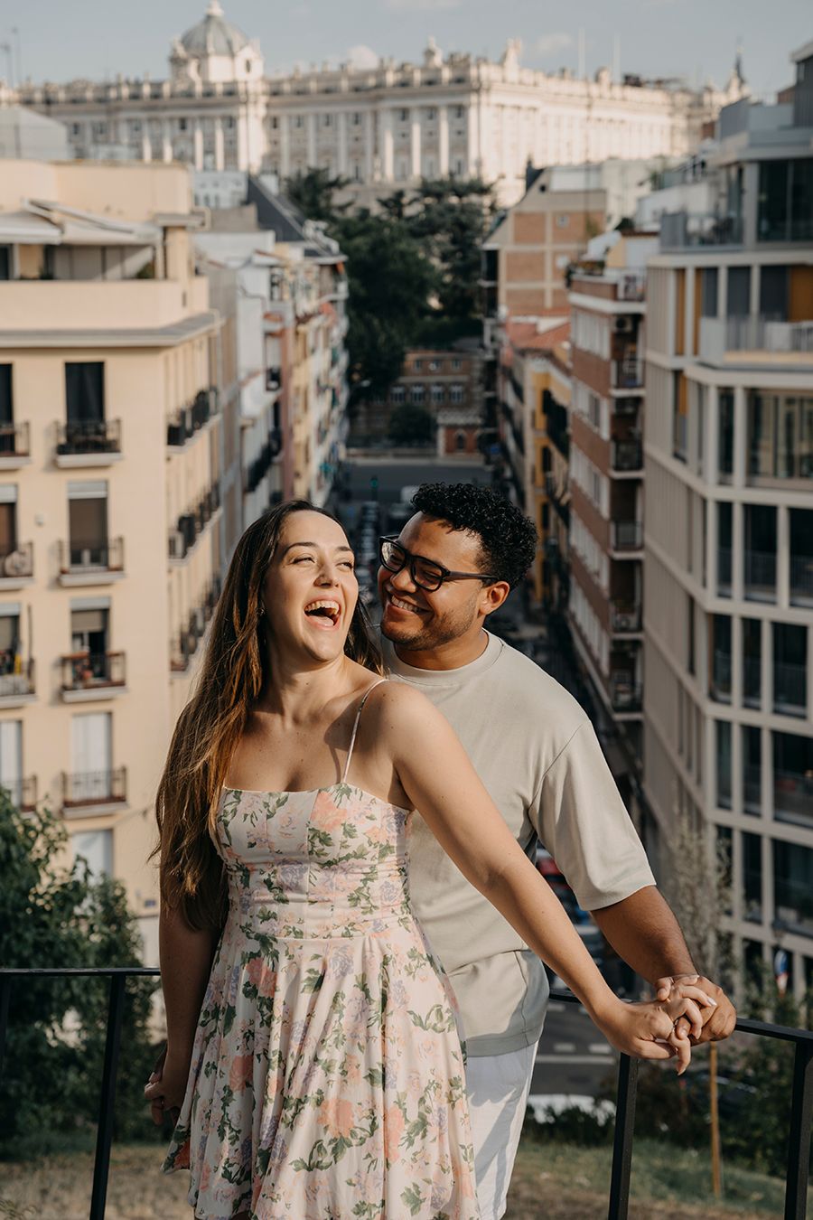 Happy couple holding hands on a balcony overlooking Madrid city streets with historic buildings in the background.
