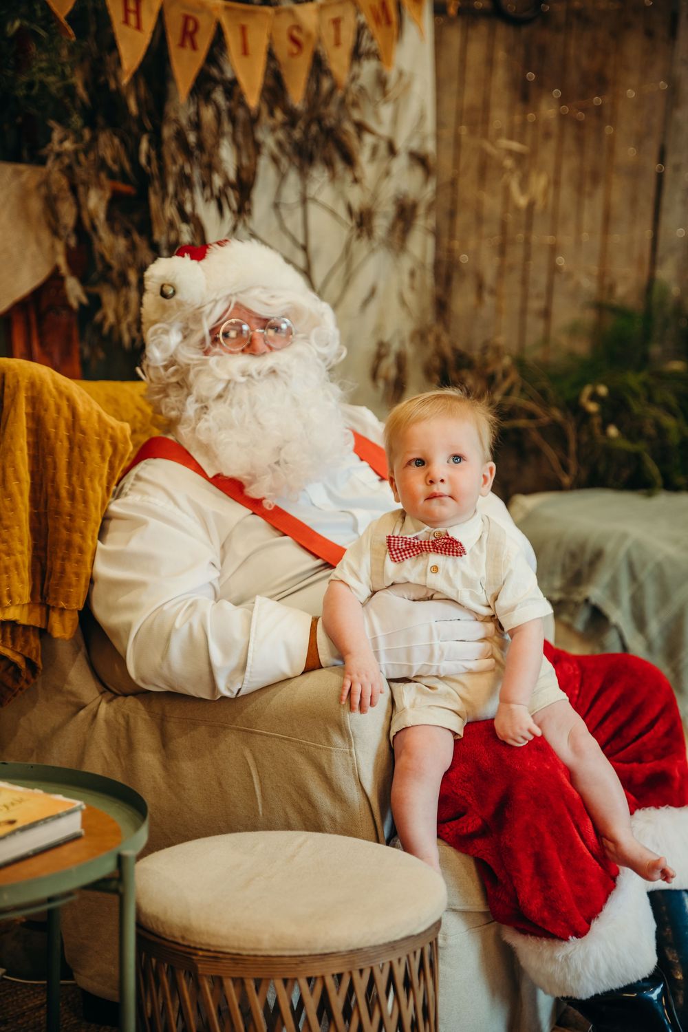 A cute baby in a cream outfit with red bow tie sits on Santa Claus's lap for a Christmas photo.