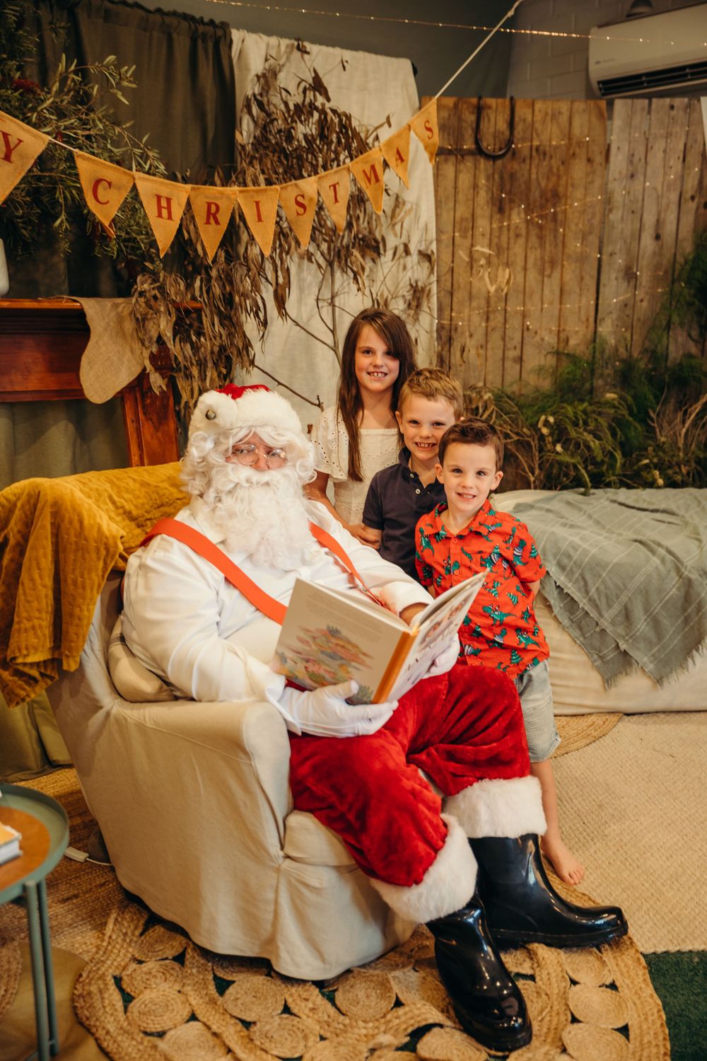 Santa Claus reads a Christmas book to three children in a cozy bush setting with Merry Christmas bunting. Kristielle Gibbons Photograpphy.