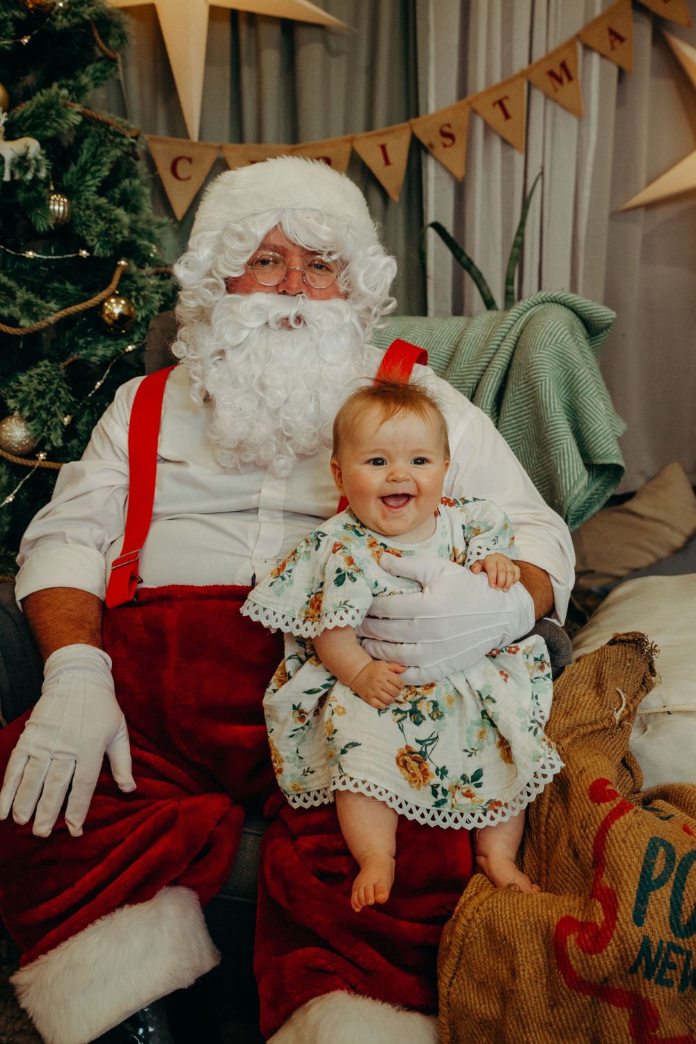 Smiling baby girl in floral dress sitting on Santa Claus lap in a festive Christmas setting.