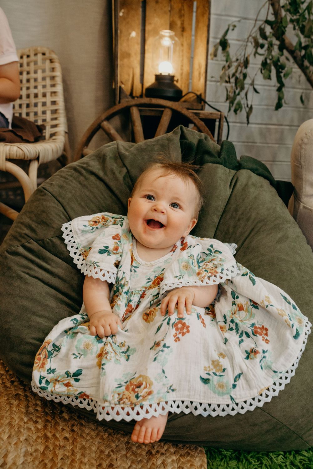 Adorable laughing baby girl in white floral dress sitting in a large green bean bag chair.