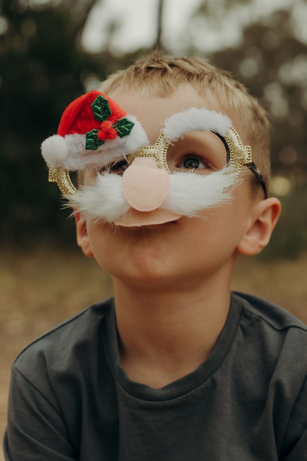 Young boy wearing festive Santa Claus novelty glasses with white beard and red hat outdoors.