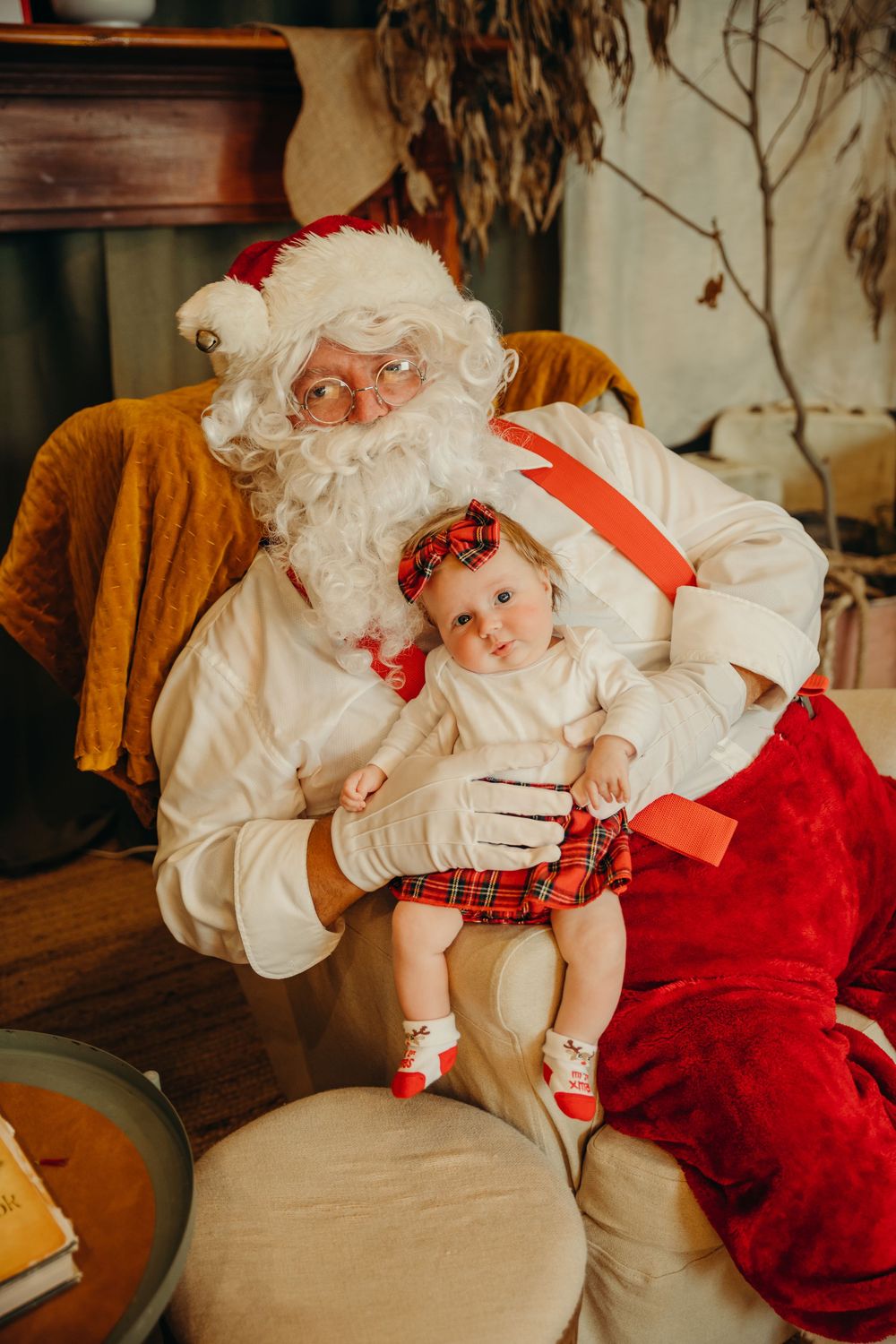 A child sits on Santa Claus's lap in a cozy bush Christmas-decorated room with a gift sack nearby. Kristielle Gibbons Photography.