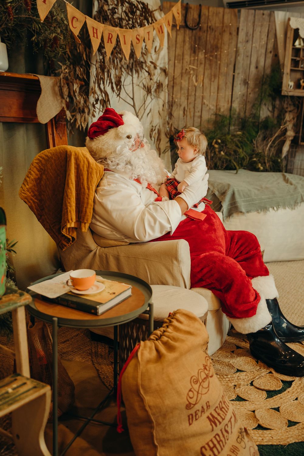 A child sits on Santa Claus's lap in a cozy bush Christmas-decorated room with a gift sack nearby. Kristielle Gibbons Photography.