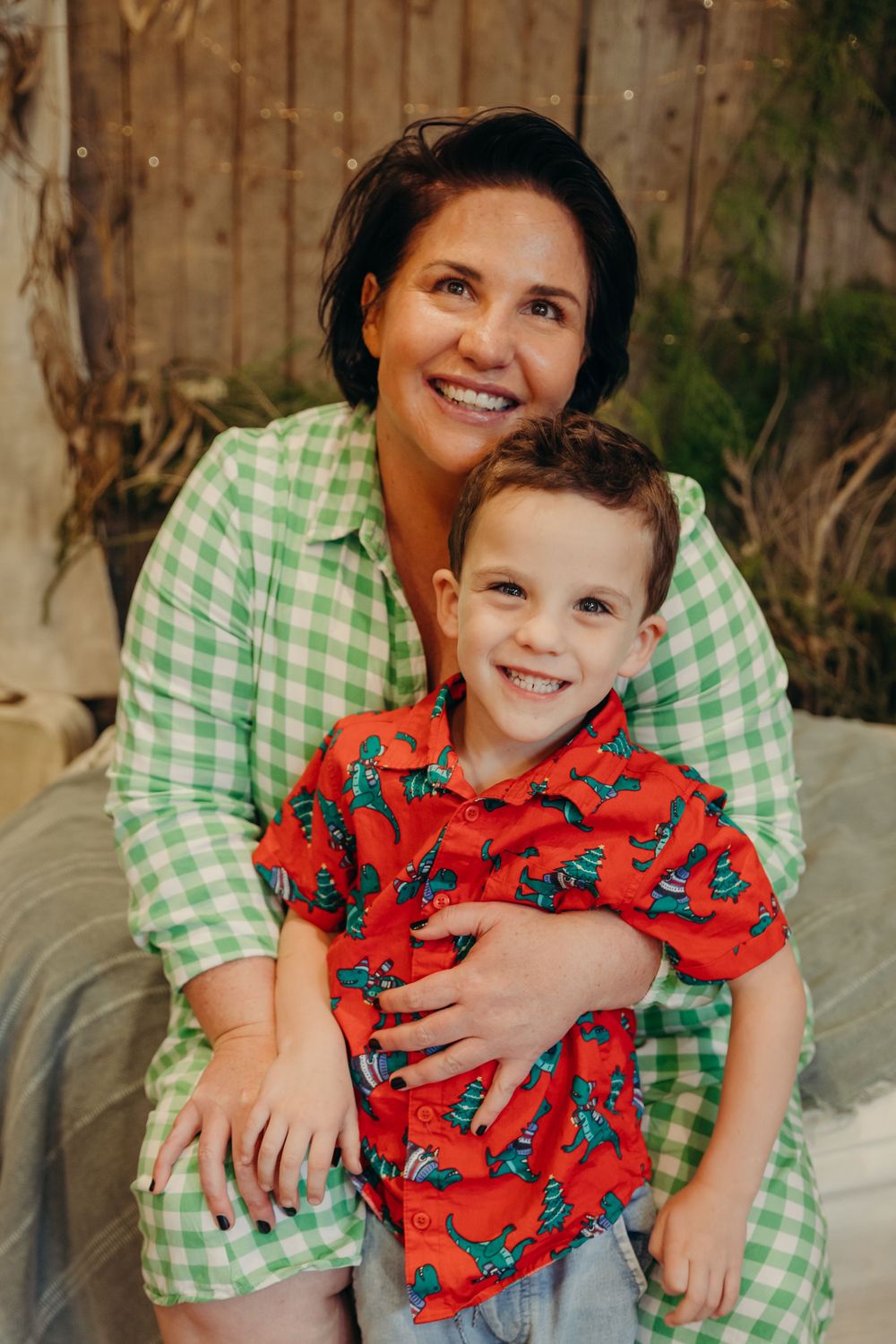 Smiling mother in green gingham dress hugs her young son in a red Christmas shirt.