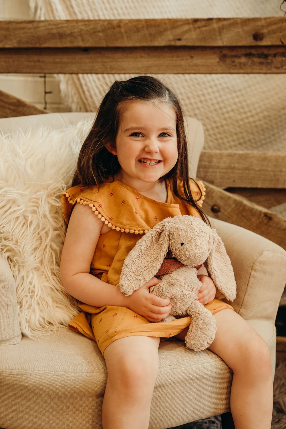 Adorable smiling toddler girl in yellow dress hugging stuffed bunny rabbit toy while sitting in cream armchair.