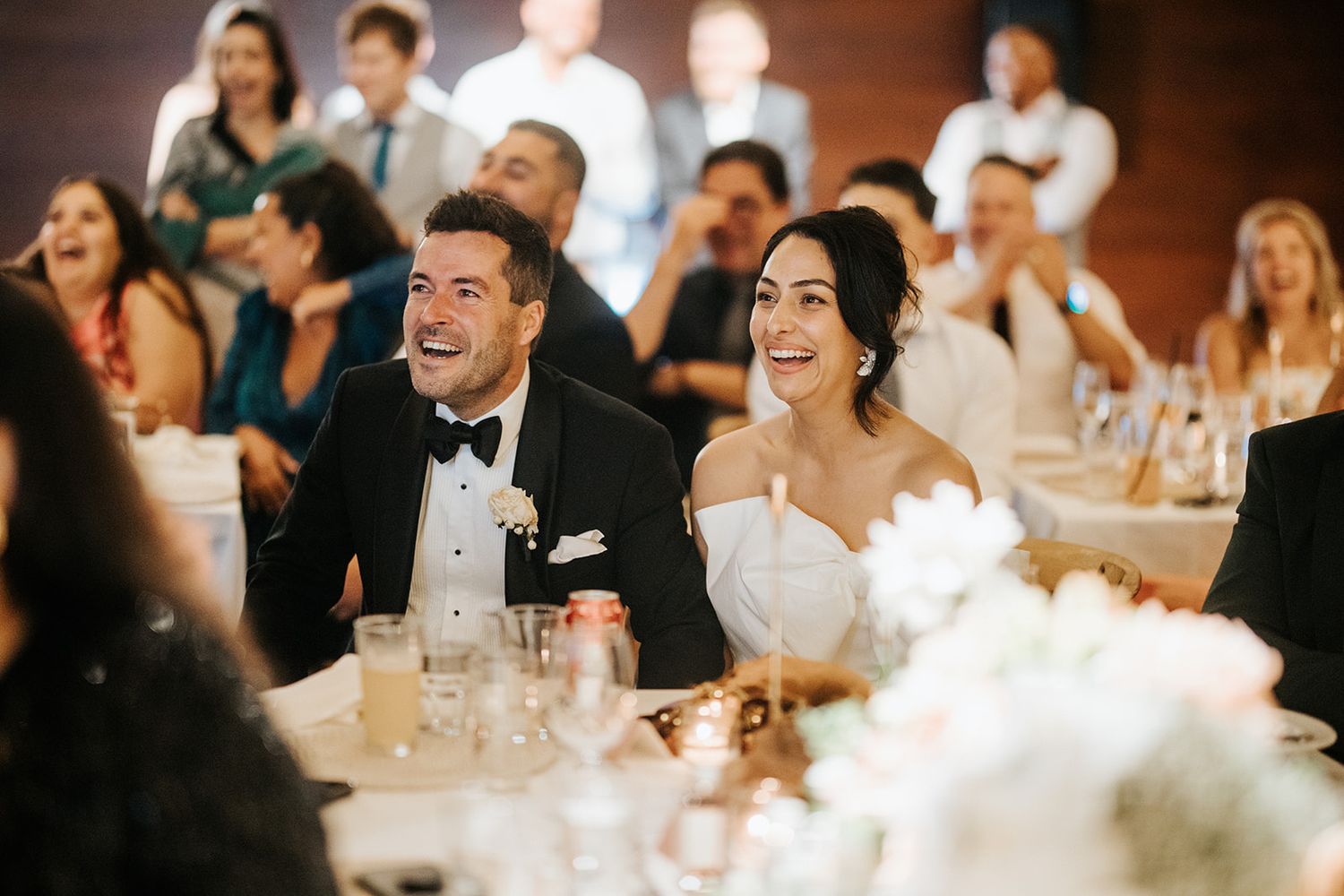 Bride and groom laughing joyfully at wedding reception table surrounded by smiling guests during speeches.