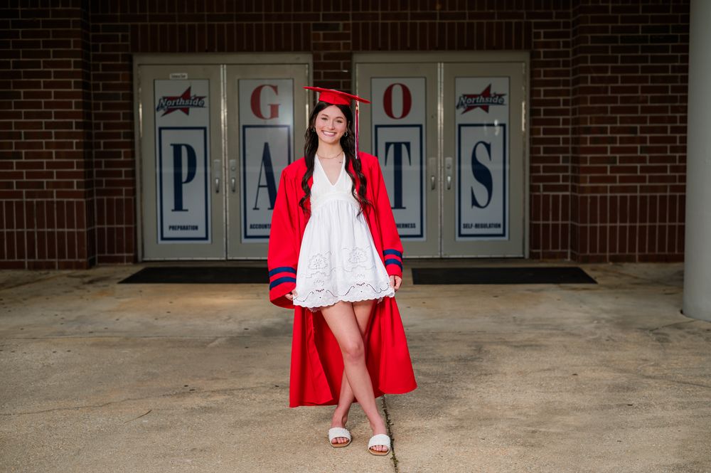 A young woman in red graduation cap and gown poses outdoors in front of a brick school building.
