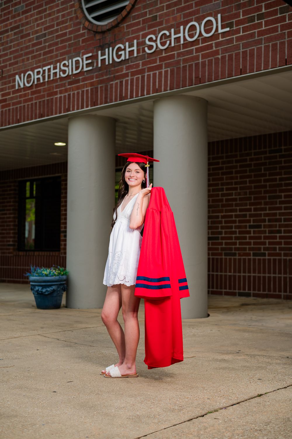 A smiling graduate holds her red cap and gown outside Northside High School wearing a white dress.