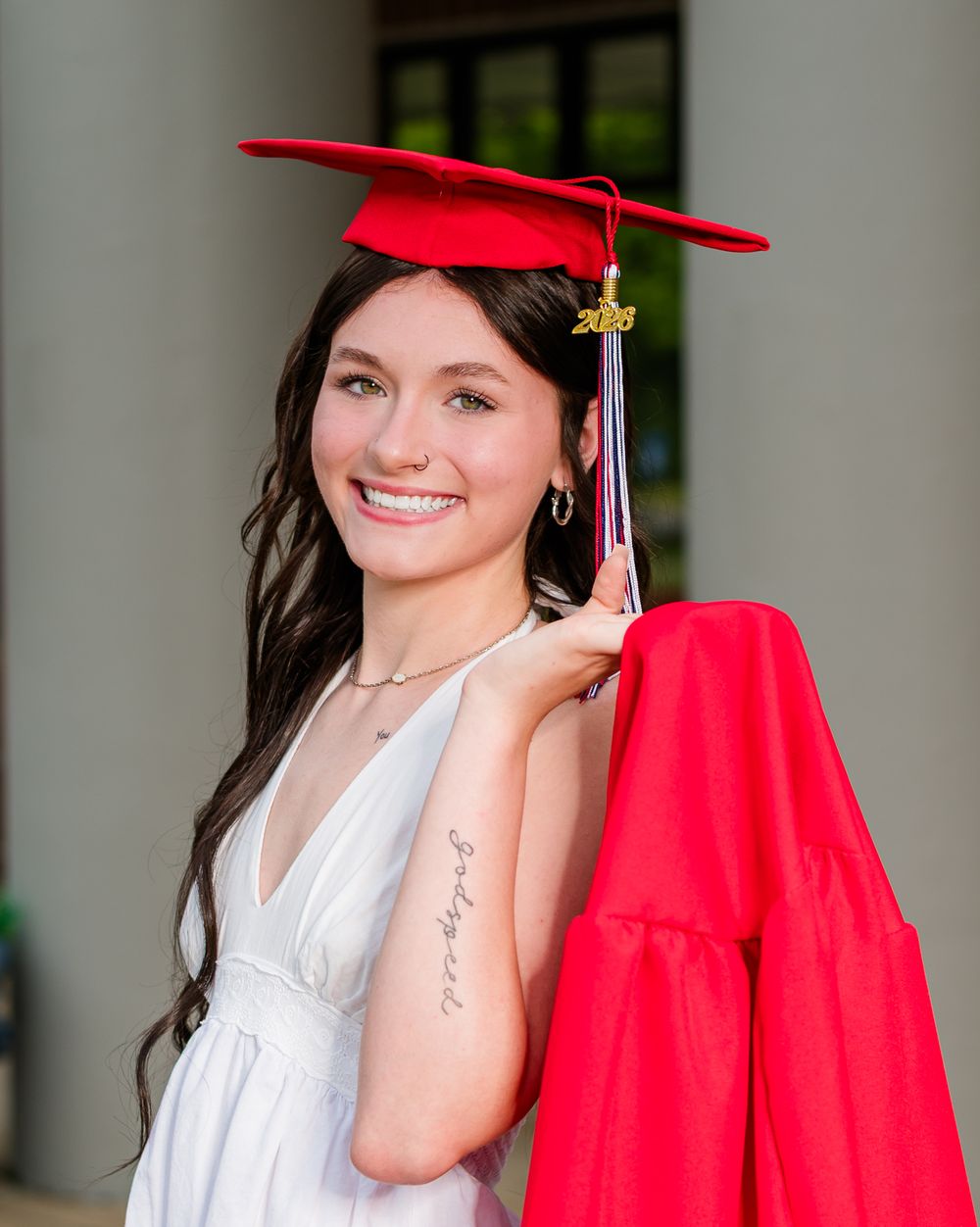 Smiling young woman in red graduation cap and gown holding diploma, wearing white dress with tattoo visible on arm.
