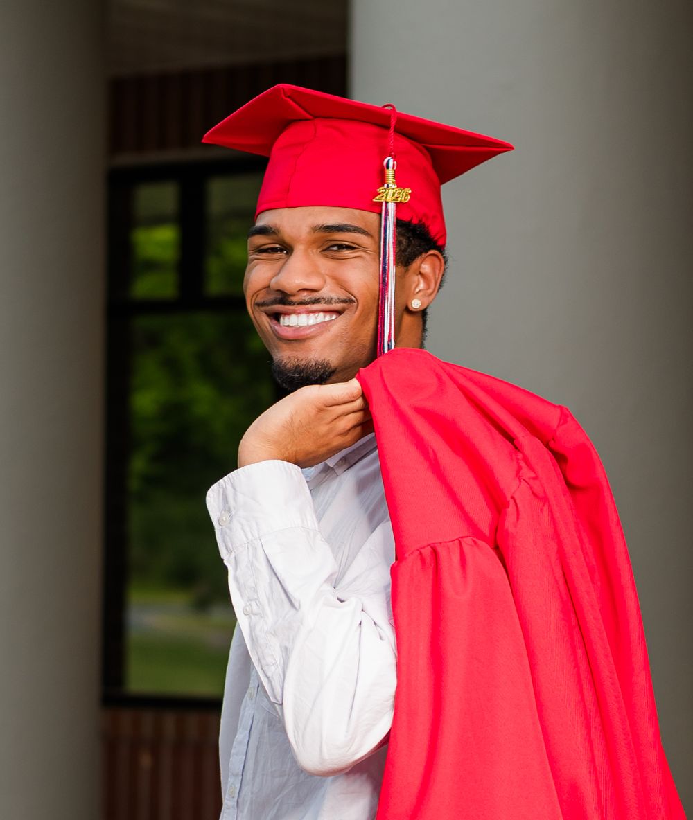 Smiling young man in red graduation cap and gown posing outdoors near white columns on graduation day.