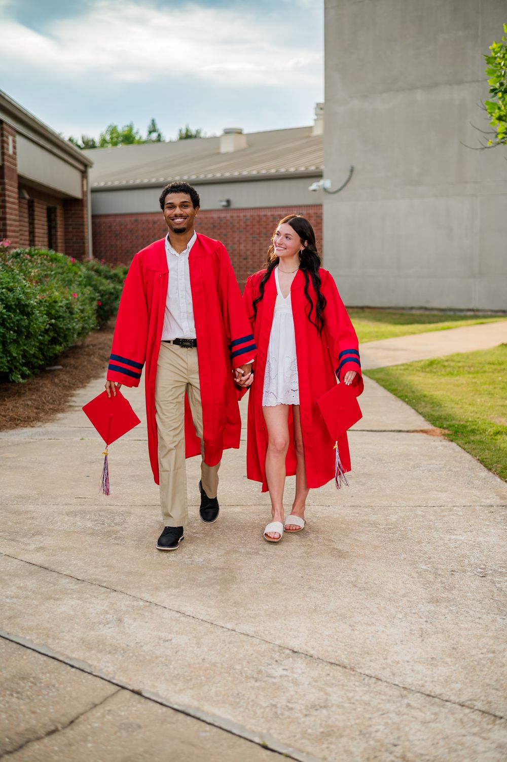 Two graduates in red robes walk hand-in-hand on a campus pathway, smiling and holding their caps.